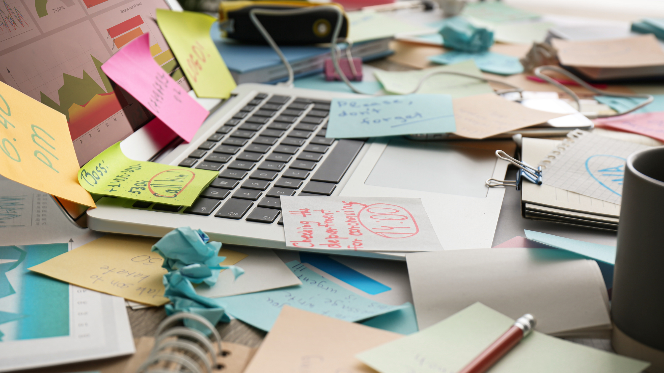 Cluttered workspace with colorful sticky notes, open laptop, paperwork, and coffee cups representing the overwhelm of managing marketing while running a small business.